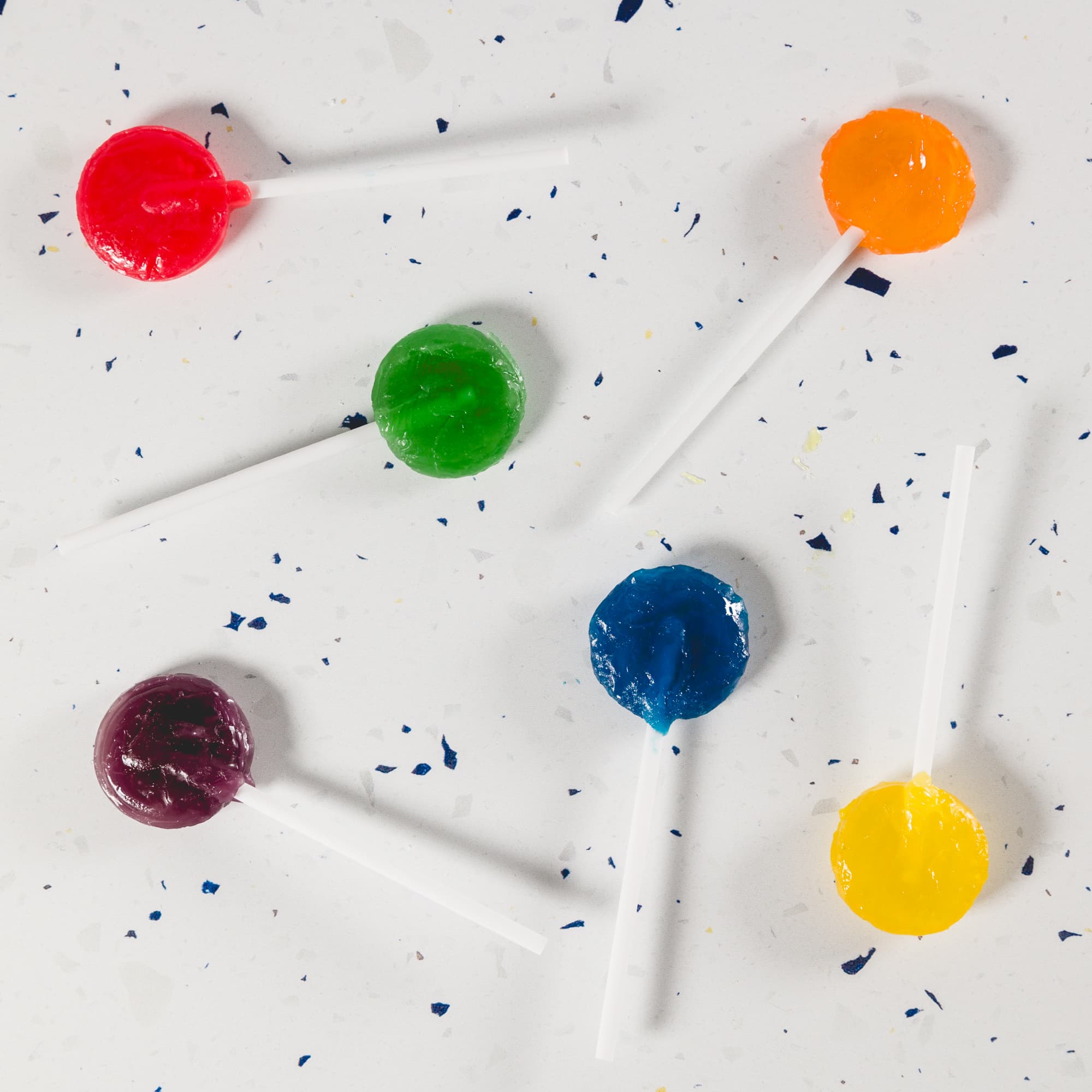 Assorted lollipops in red, green, orange, blue, yellow, and purple on a textured surface, featuring white sticks.