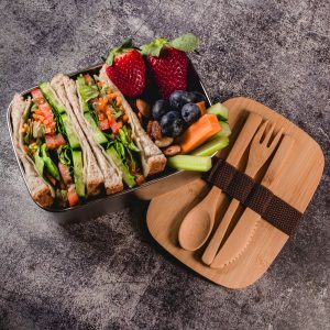 A stainless steel lunch box with a bamboo lid. Contains wraps, fruits, and utensils. Features a logo.
