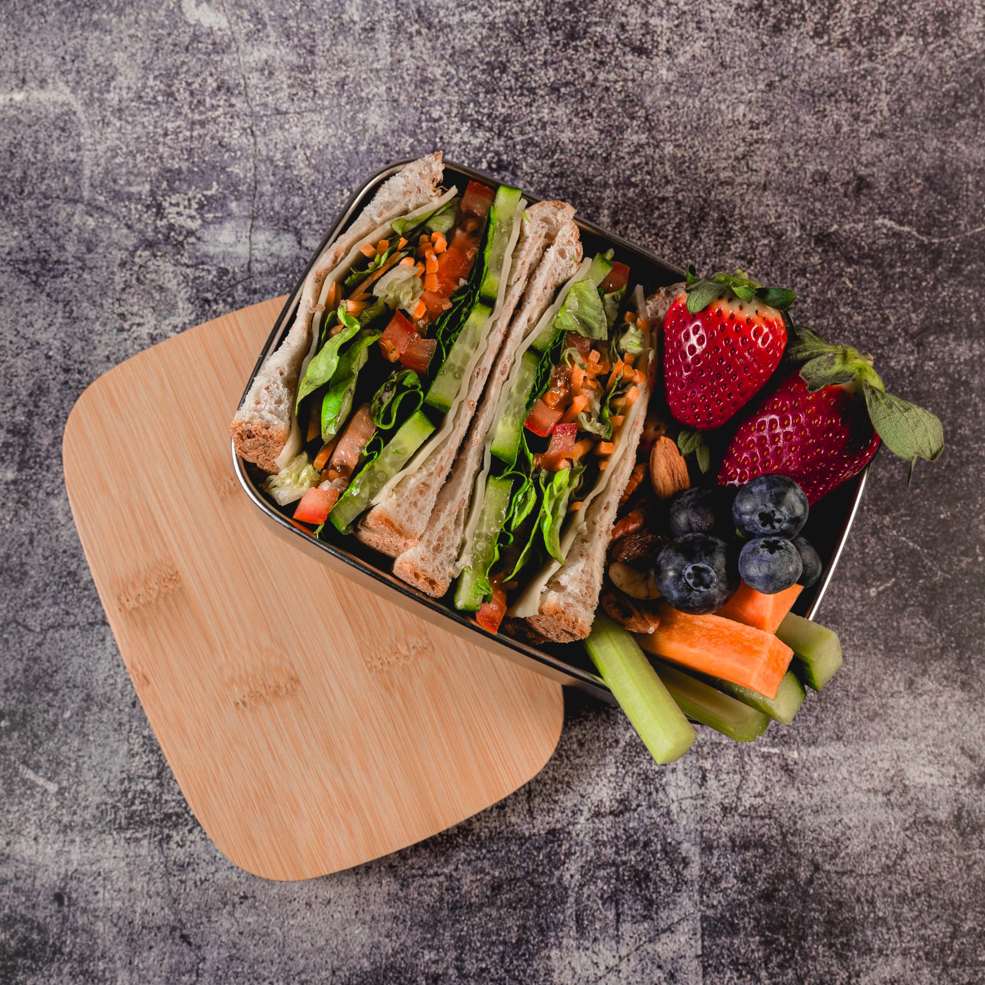 A lunch box filled with two sandwiches, strawberries, blueberries, celery, and carrots, with a wooden lid.