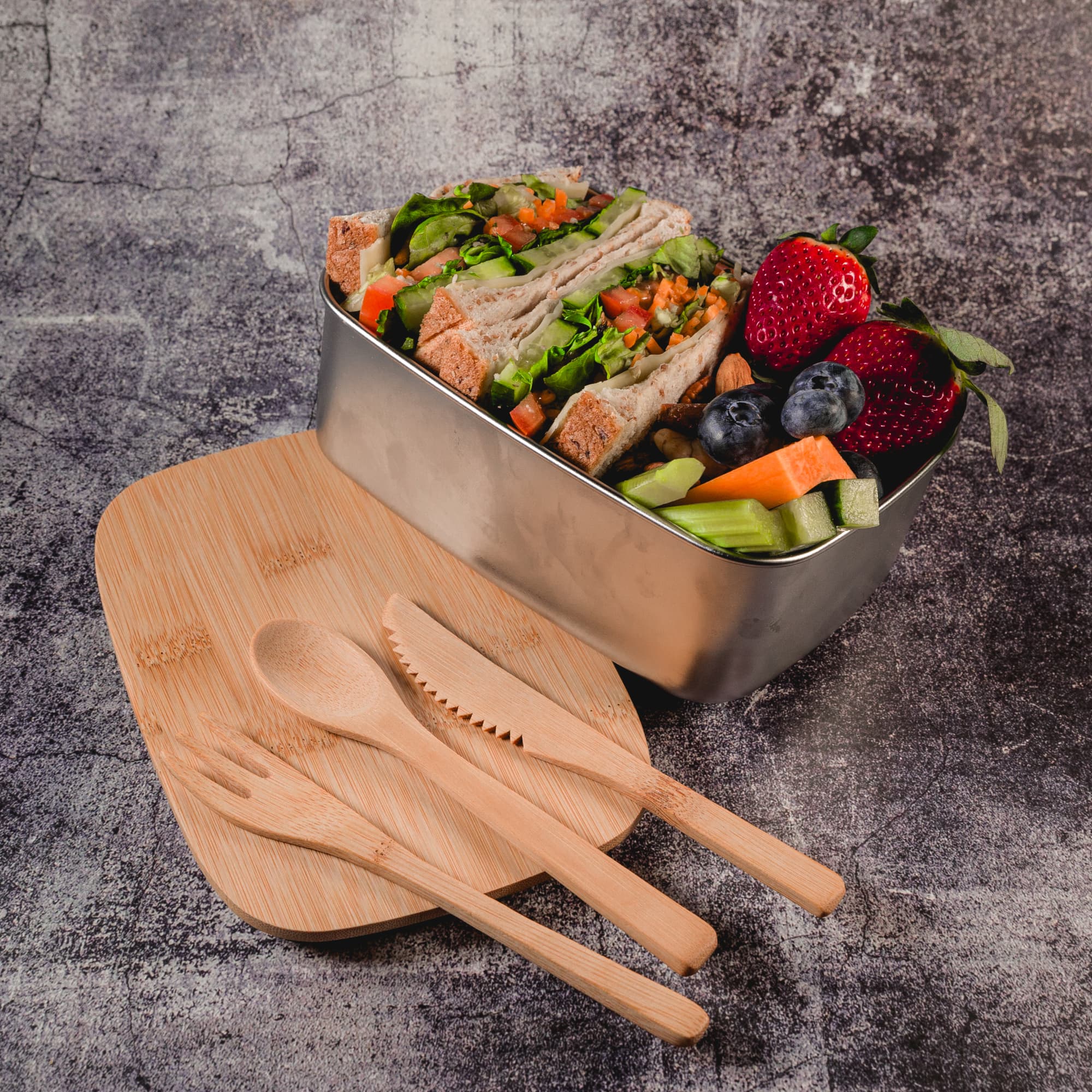 A stainless steel lunch box filled with sandwiches, mixed fruits, and vegetables, accompanied by wooden utensils.