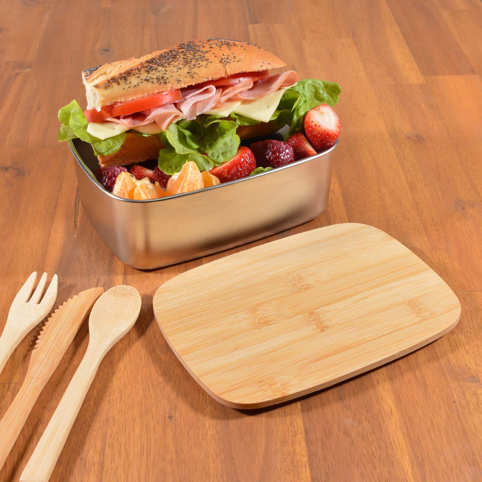 A metal lunch box filled with a sandwich, lettuce, strawberries, and a cutting board made of wood beside it.