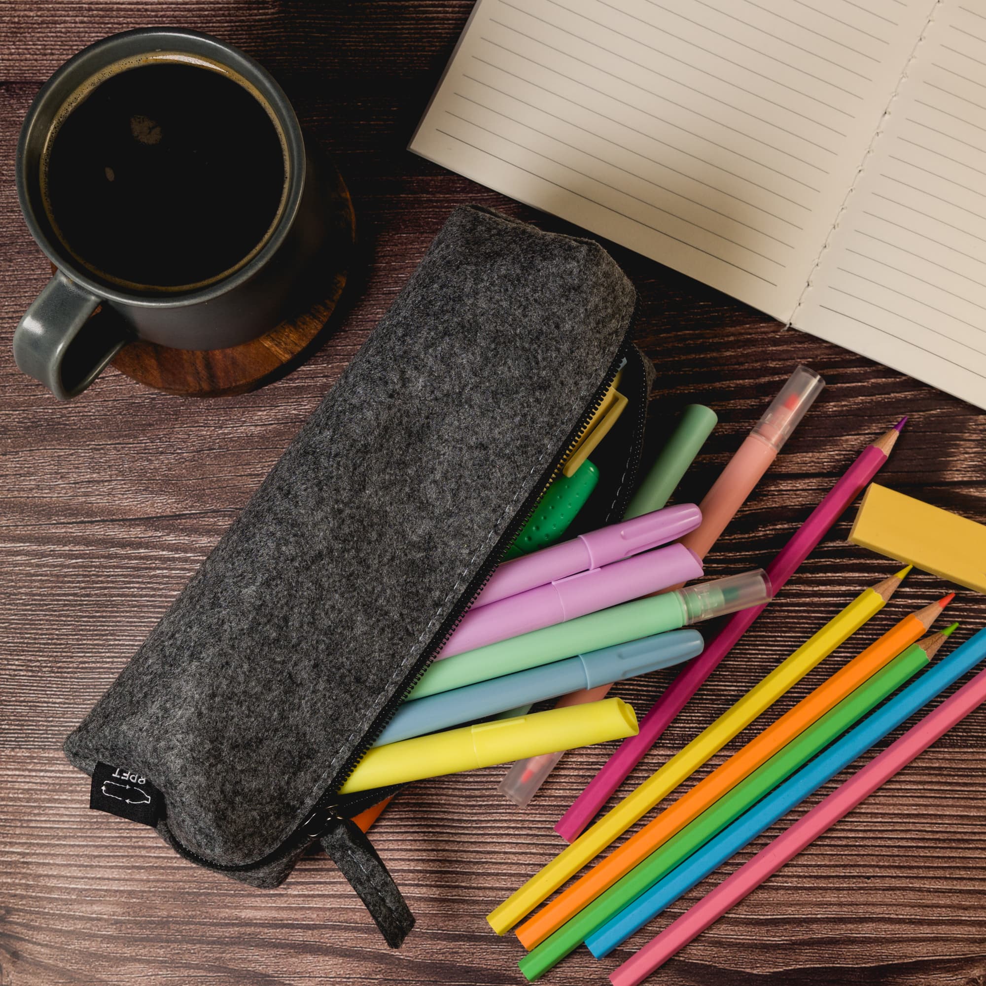A gray felt utility case surrounded by colorful pens, with a coffee mug and notebook on a wooden surface.