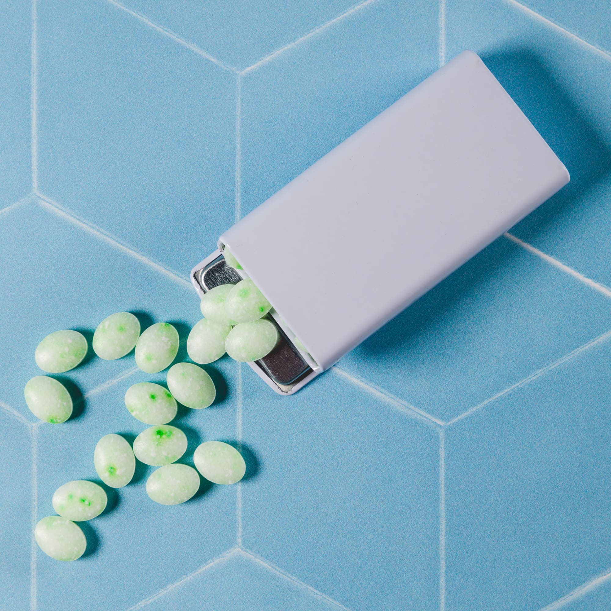 A flip top tin containing spearmint mints, mint green in color, with some mints scattered on a blue tiled surface.