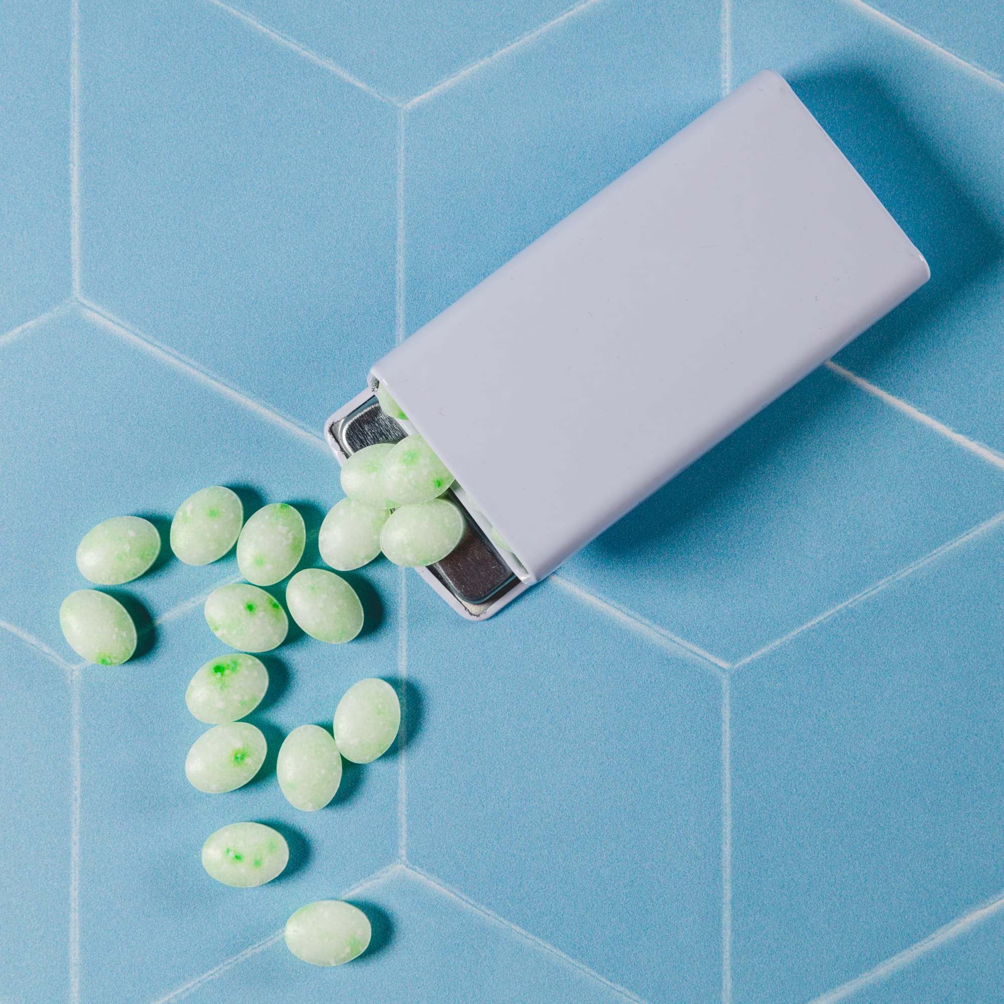 A flip-top tin containing green spearmint candies, with some candies spilled out on a blue hexagonal surface.