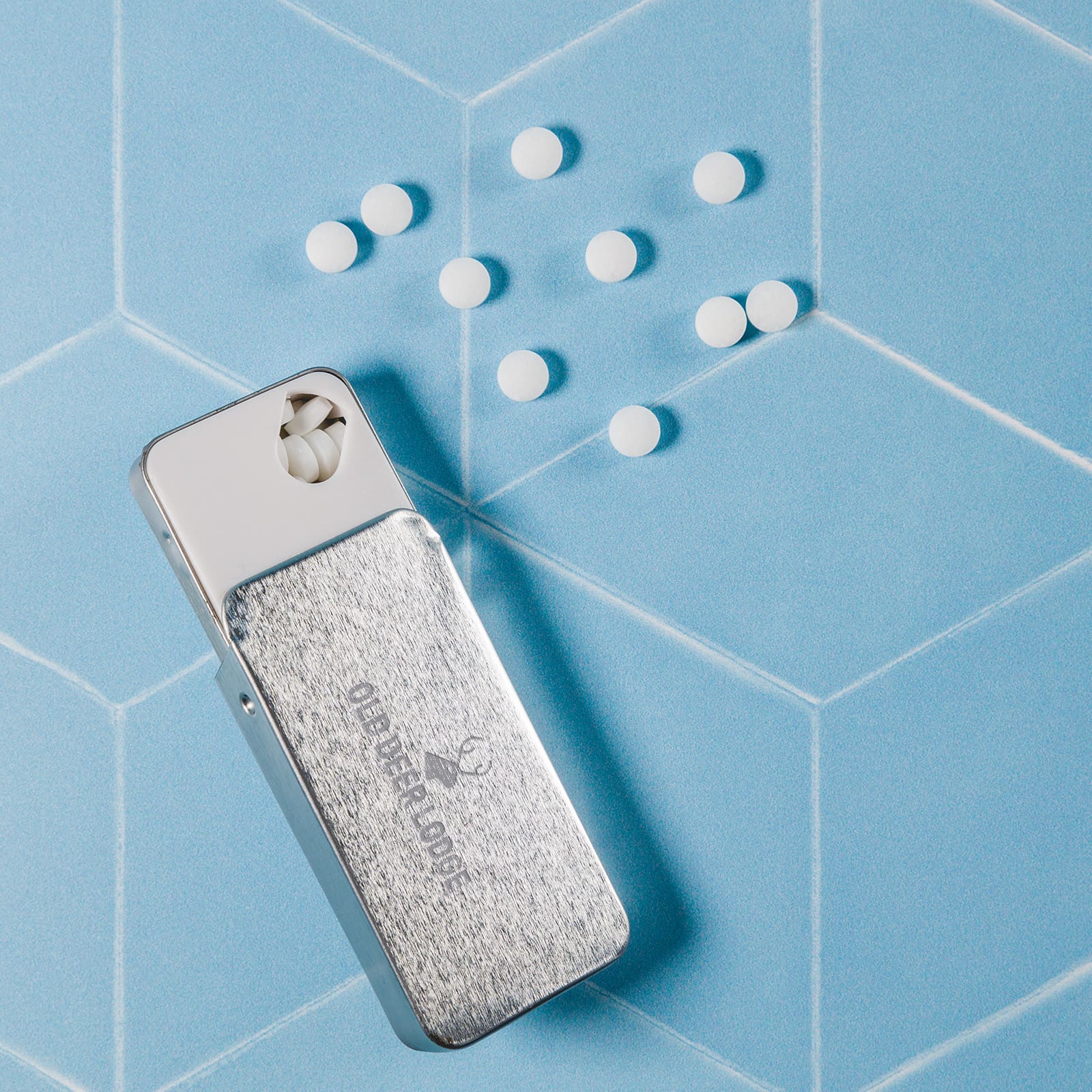 A silver slider tin containing sugar-free breath mints, with white mints scattered beside it on a pastel blue background.