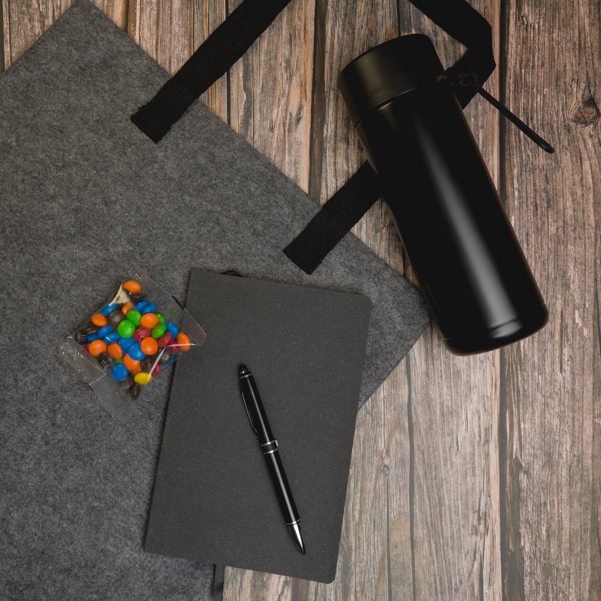 A black drink bottle, a black notebook, a pen, and a clear container of colorful candies on a grey tote bag.