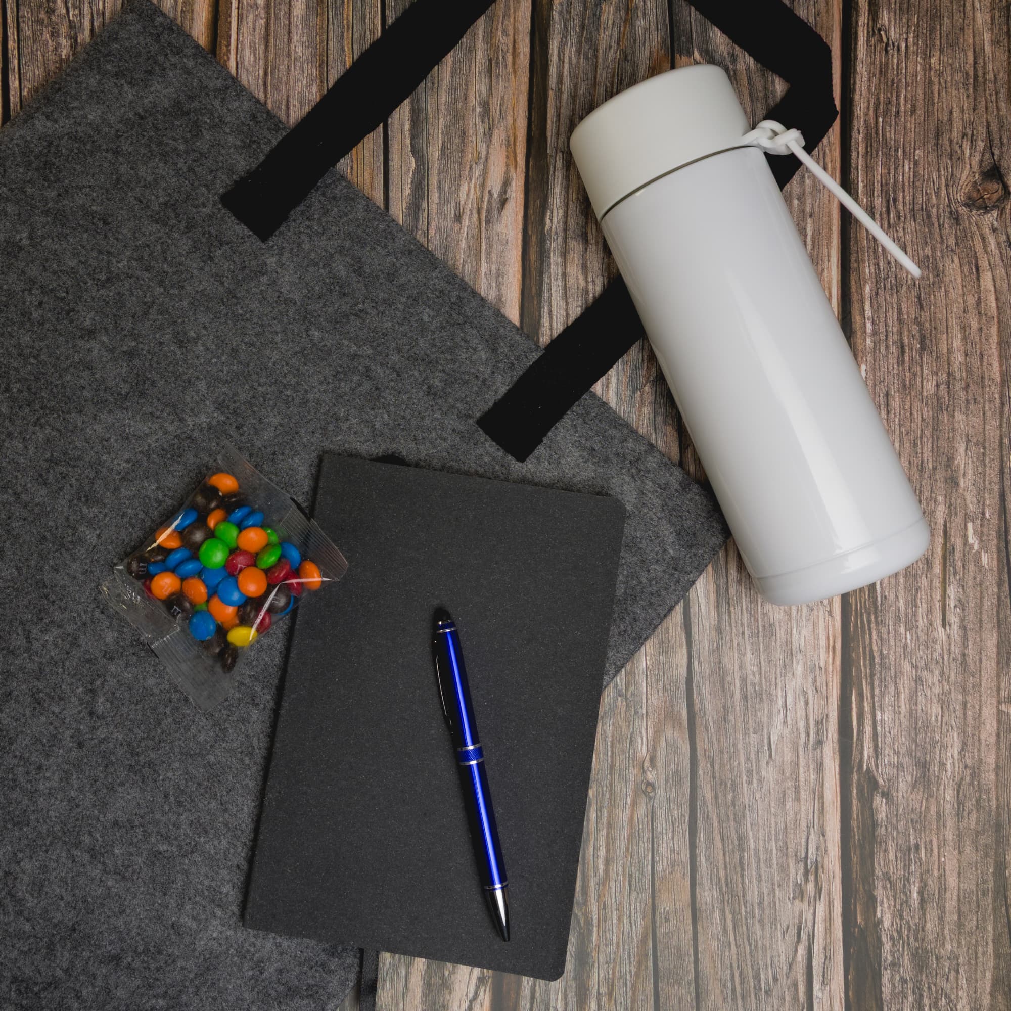 A gray tote bag, a white drink bottle, a black notebook, a blue pen, and a clear container filled with colorful candies.