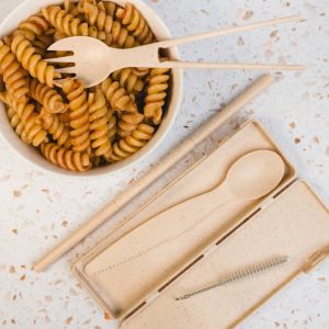 A tan Ramen Cutlery Set with a spoon, fork, and chopsticks, placed beside a bowl of rotini pasta.