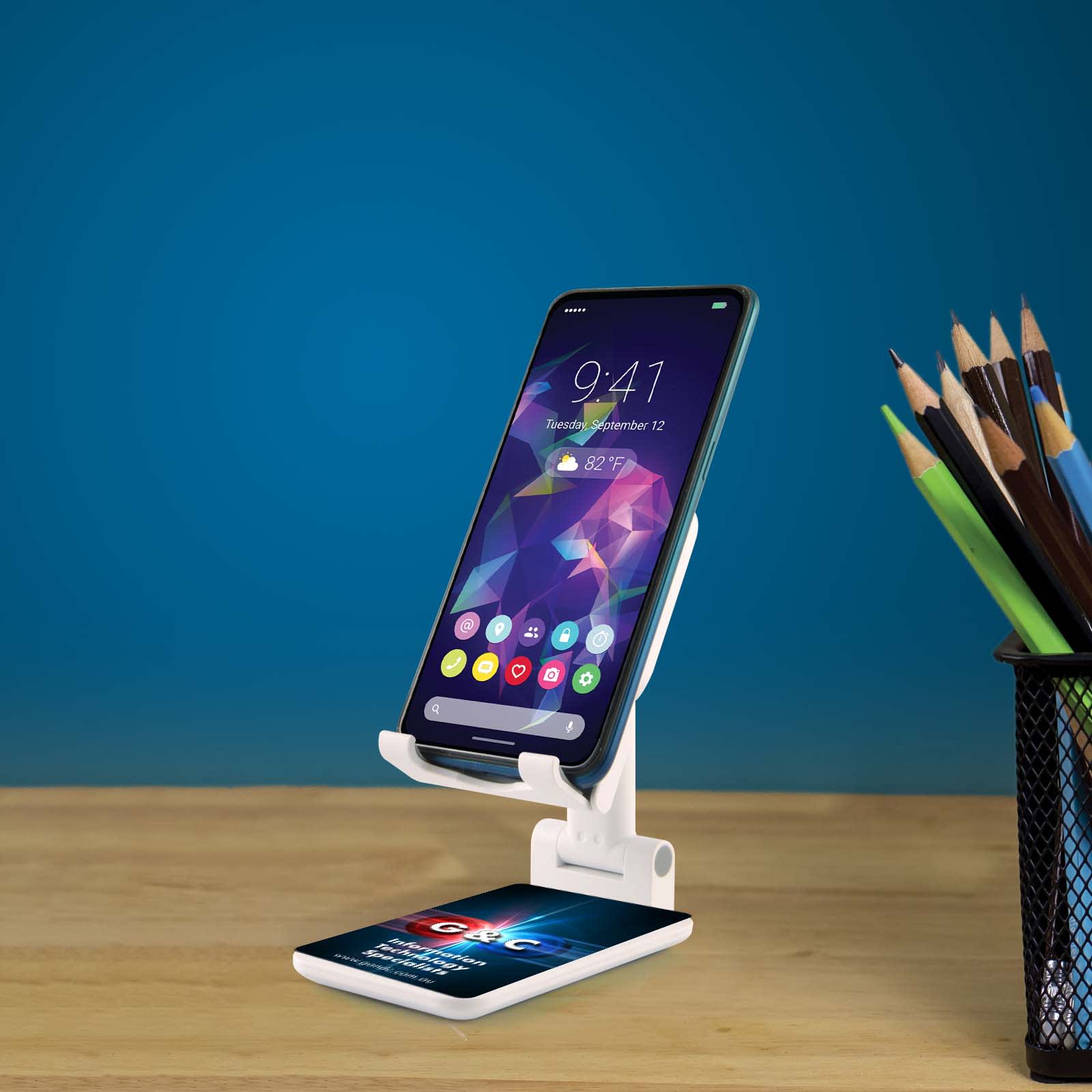 A white phone stand holds a smartphone, placed on a wooden table beside a holder of colored pencils against a blue background.
