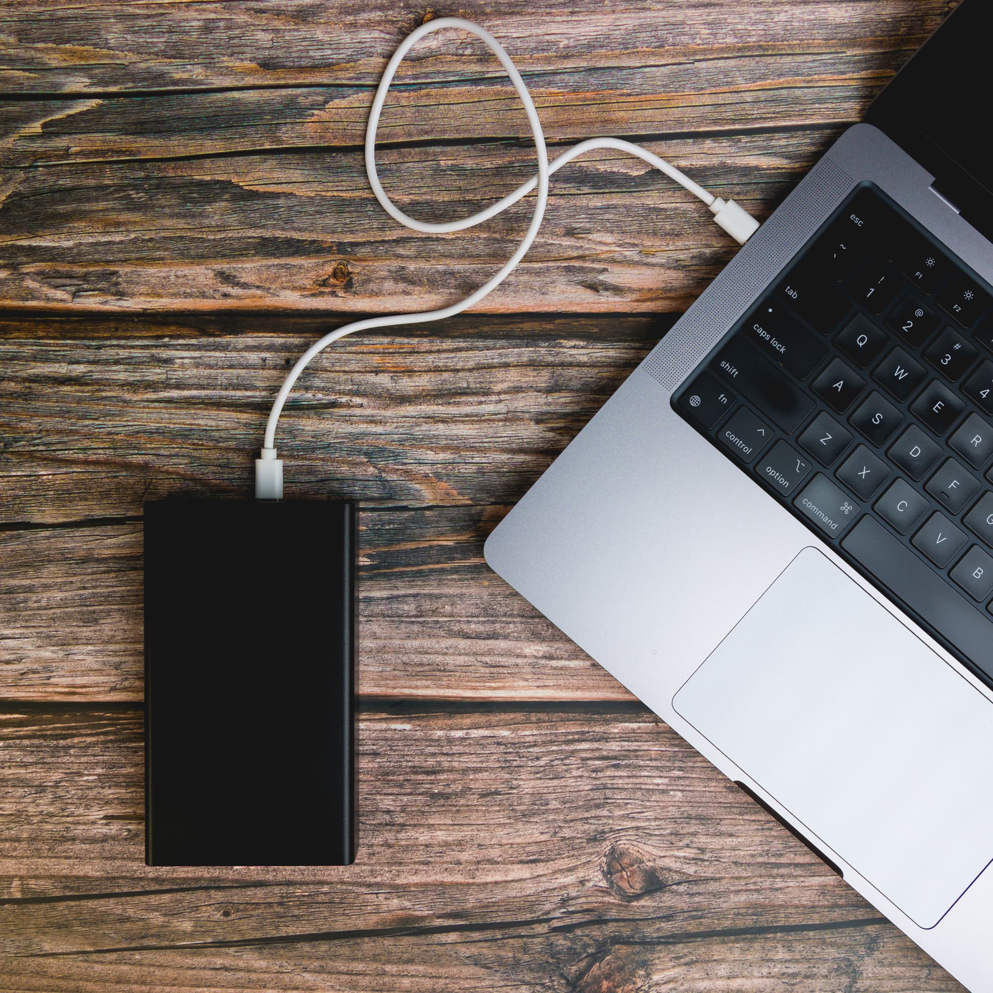 A power bank named Ortega is connected to a laptop via a charging cable, set against a rustic wooden surface.