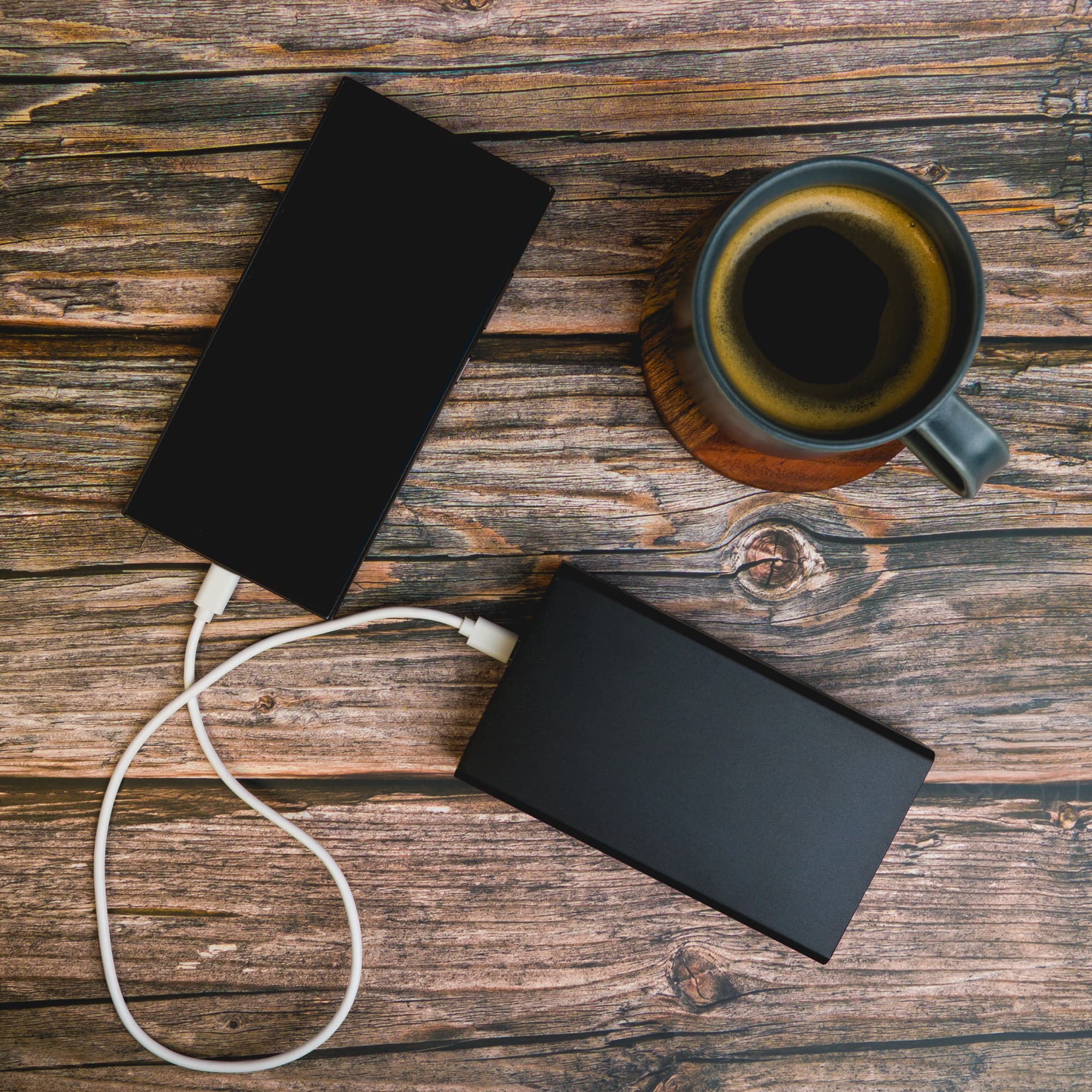 Two black power banks with white charging cables, placed alongside a cup of coffee on a wooden surface.