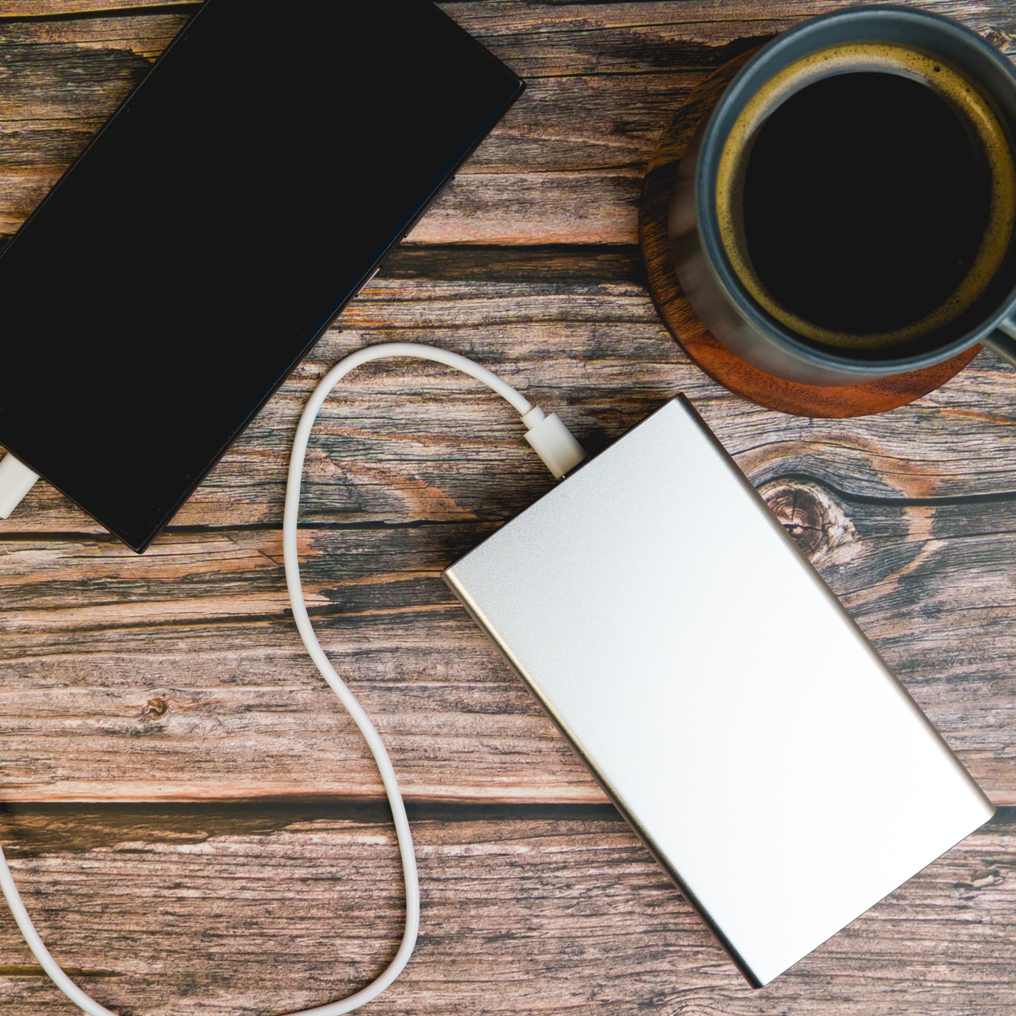 A silver and a black power bank are connected by a white cable, placed on a wooden surface beside a coffee cup.