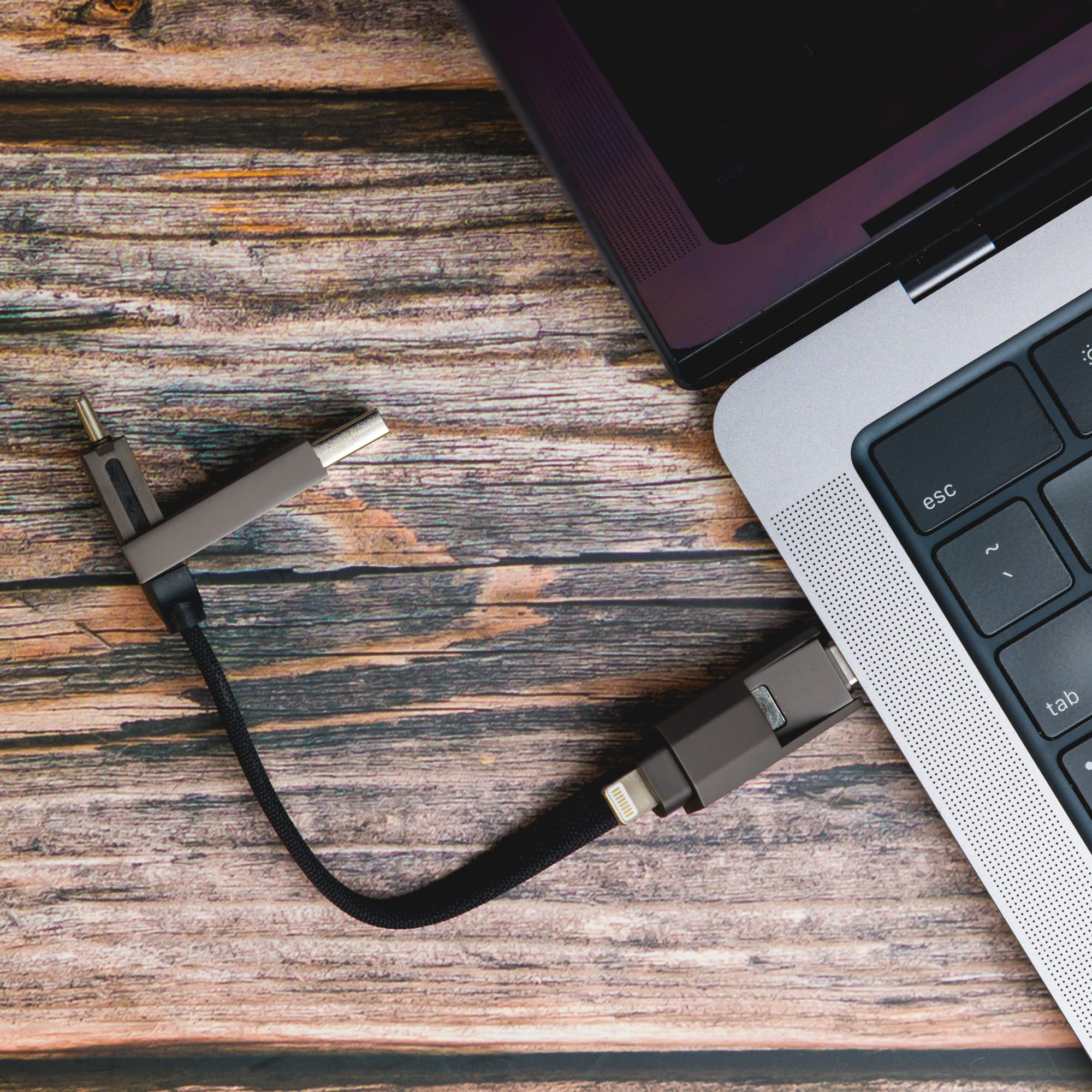 A 4-in-1 charging cable in gray, connected to a laptop, resting on a wooden surface. The cable features multiple connectors.