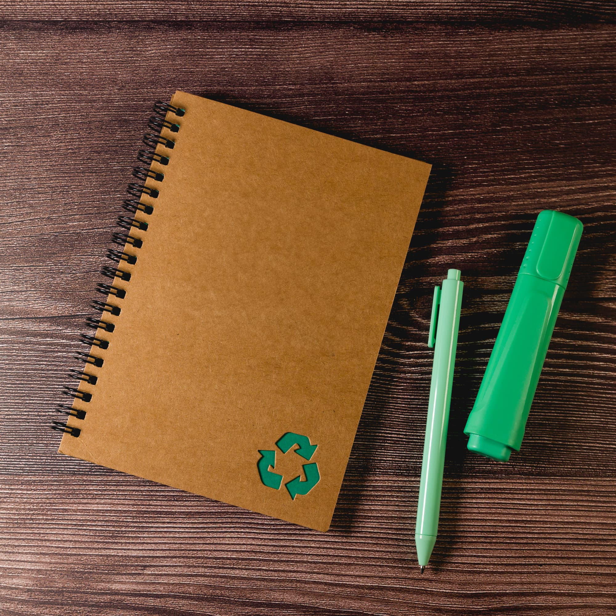 A brown spiral notebook with a recycling symbol, accompanied by a green pen and a green highlighter.