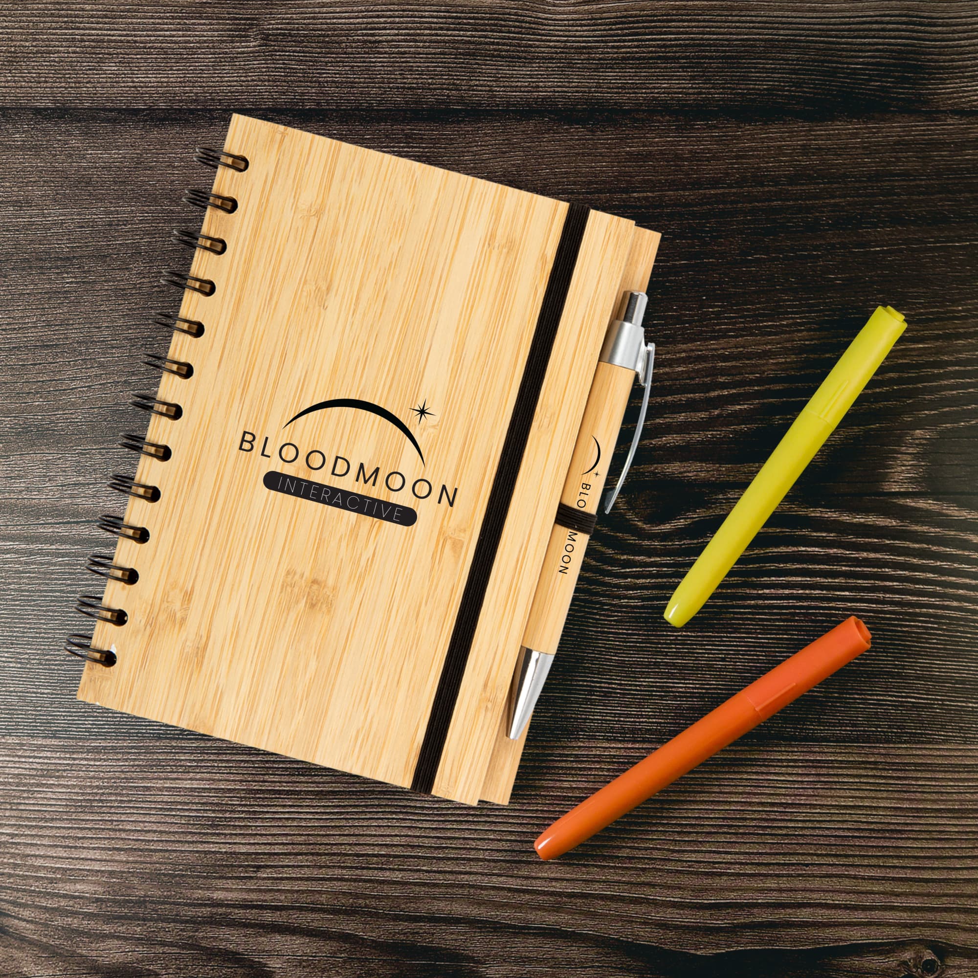 A bamboo notebook with a spiral binding, accompanied by a pen and three colored markers: yellow, orange, and green.