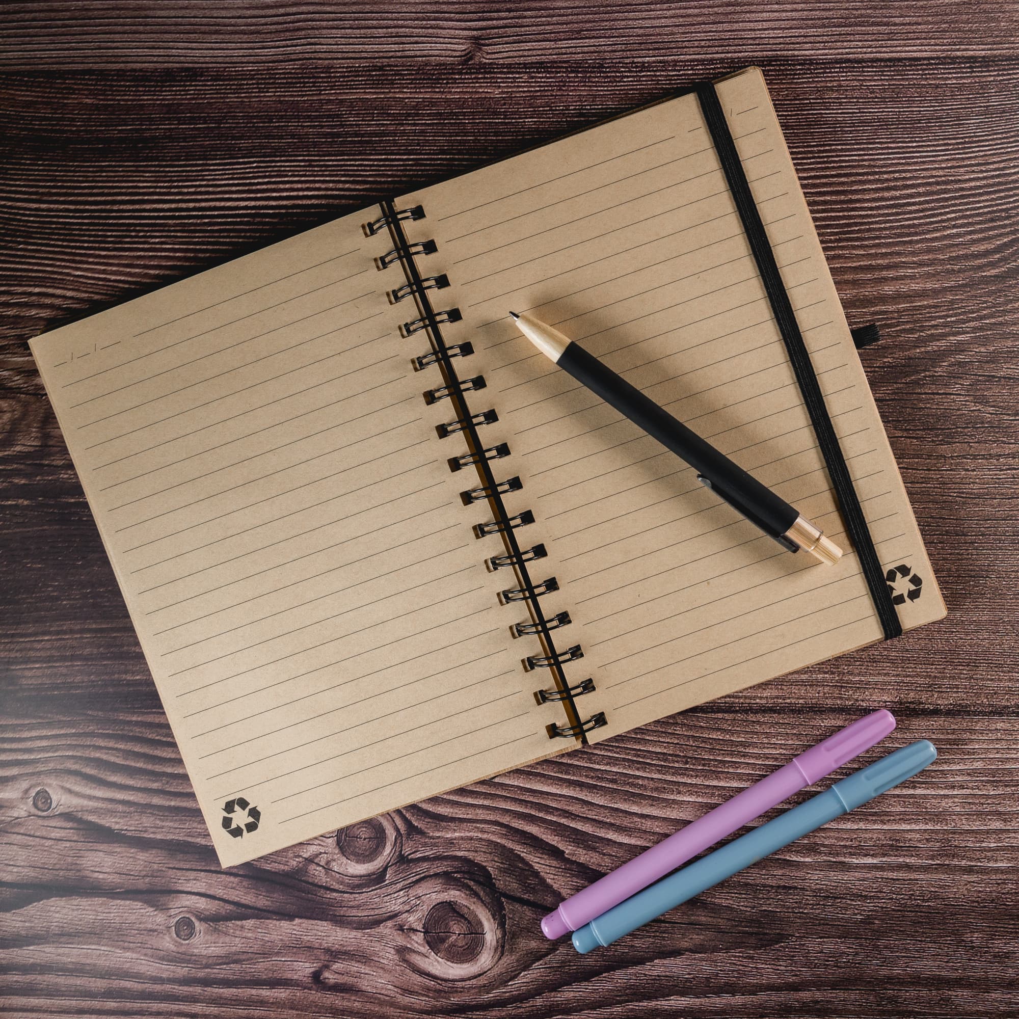 A spiral A5 notebook with a brown cover, closed with a black pen and colorful markers beside it.