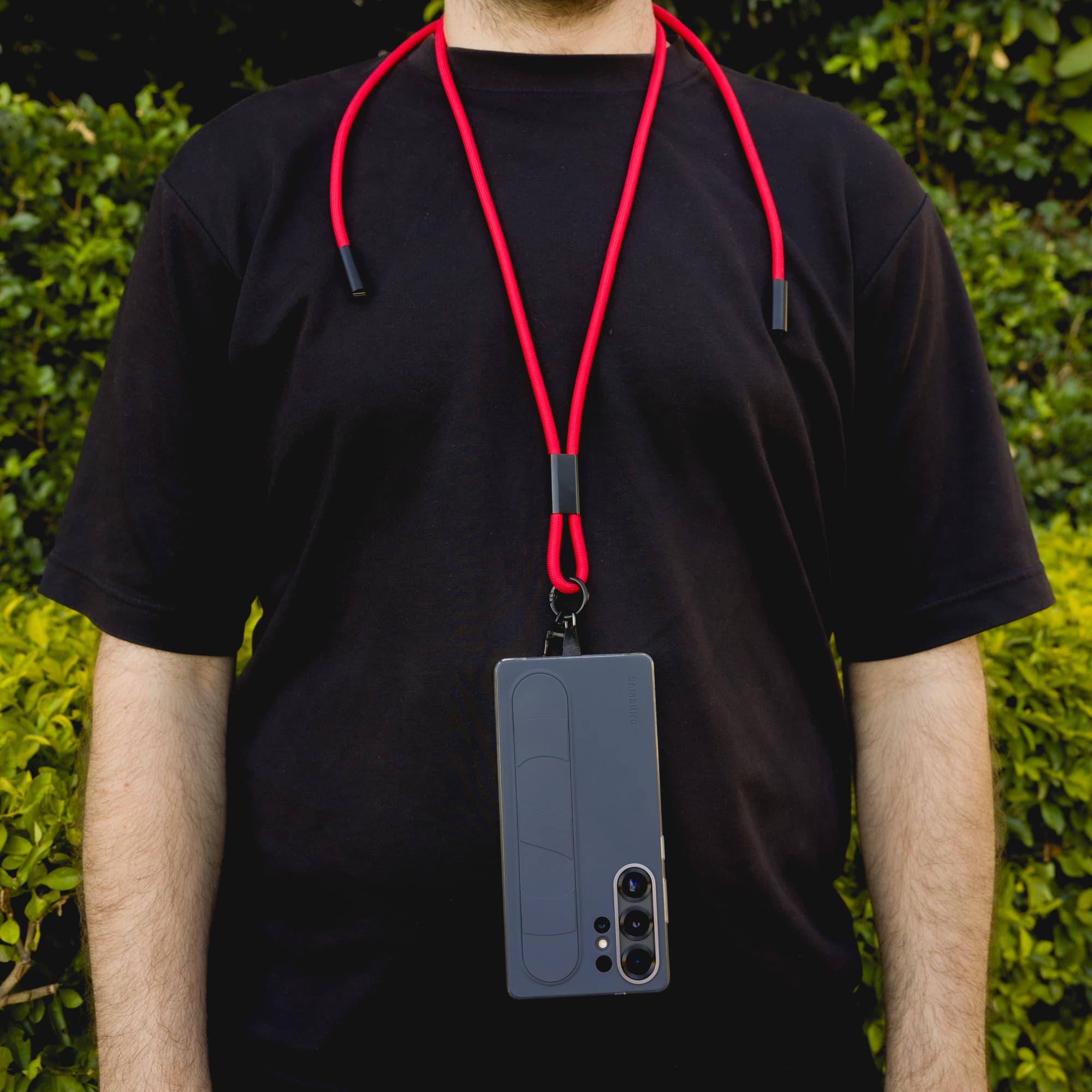 A phone lanyard in red, holding a smartphone, worn around a person's neck against a green backdrop.
