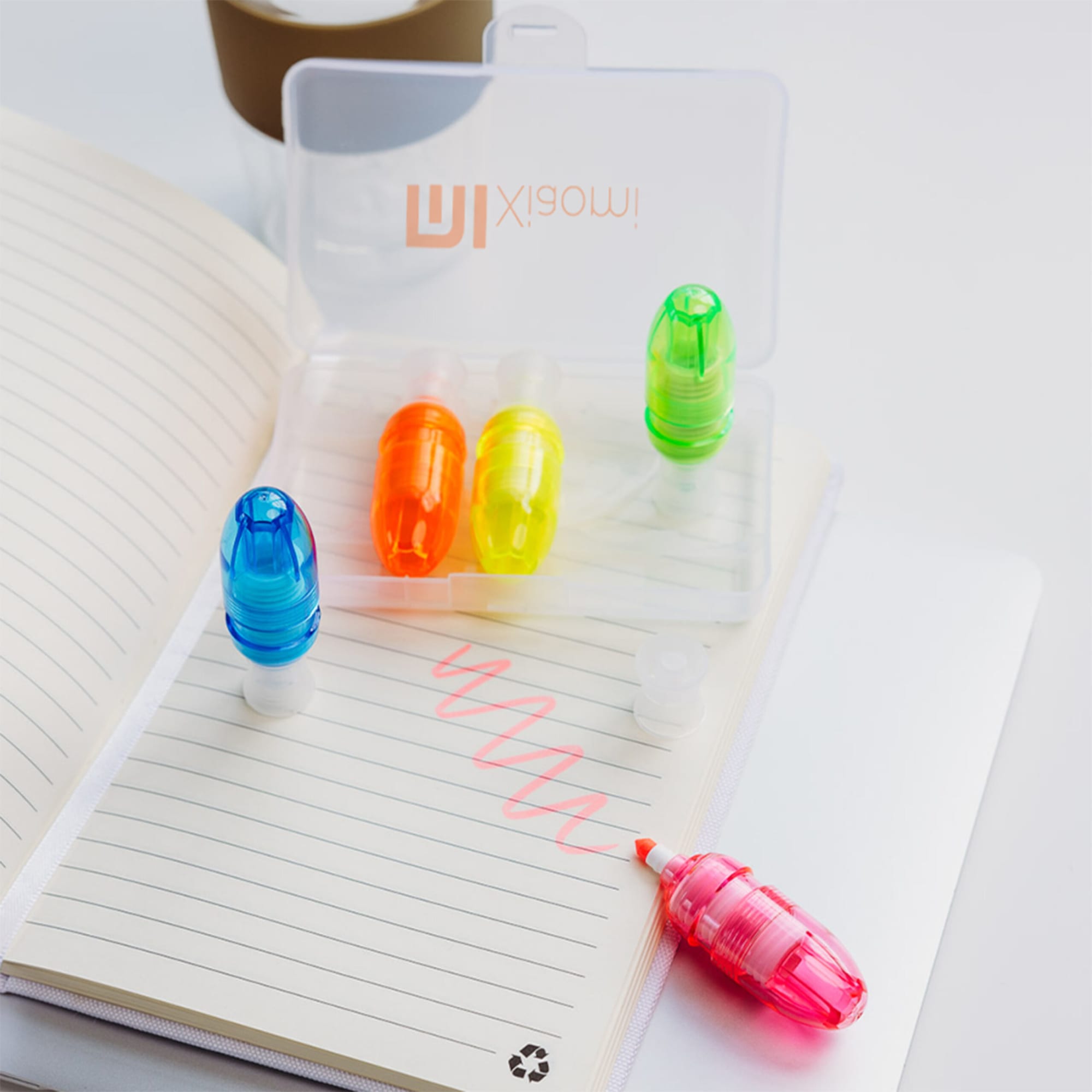 A transparent case containing four highlighters in blue, orange, yellow, and pink, placed on a notepad with lines.
