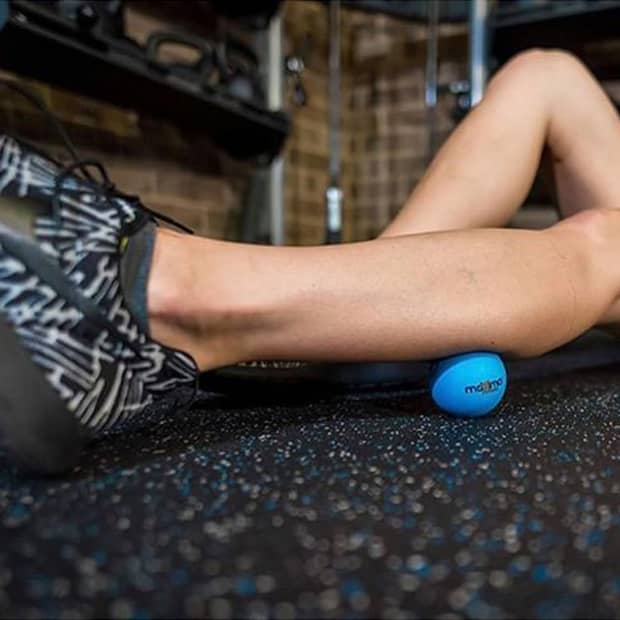 A blue branded massage ball rests on a gym floor, with a person's leg applying pressure on it.