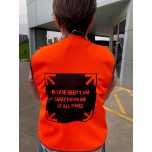 An orange safety vest with a black message graphic on the back instructing to maintain a distance of 1.5 metres.