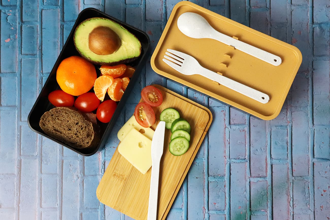 A two-layer lunch box in black and beige contains utensils and food items like fruit, bread, cheese, and vegetables.