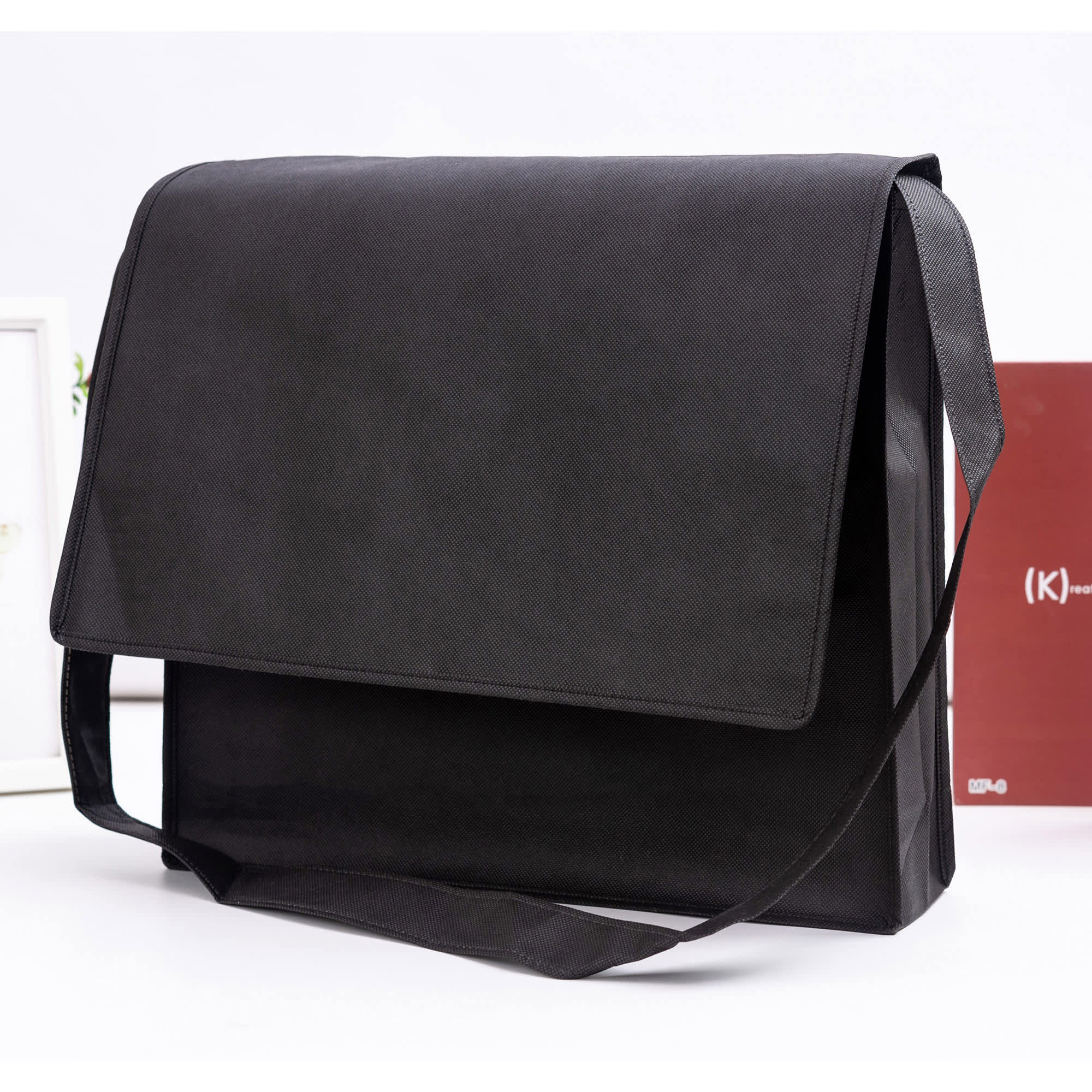 A black non-woven flap satchel with a shoulder strap and branding logo displayed.