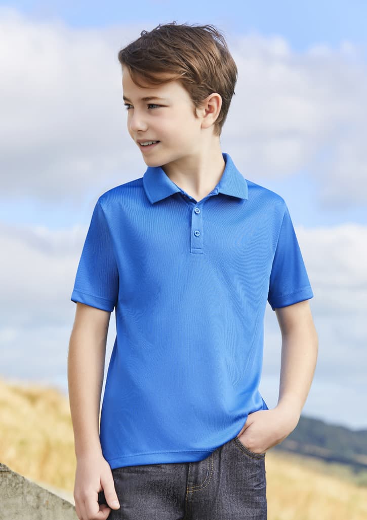 A blue short sleeve polo shirt on a young boy, standing outdoors with a scenic background. The shirt has a logo.