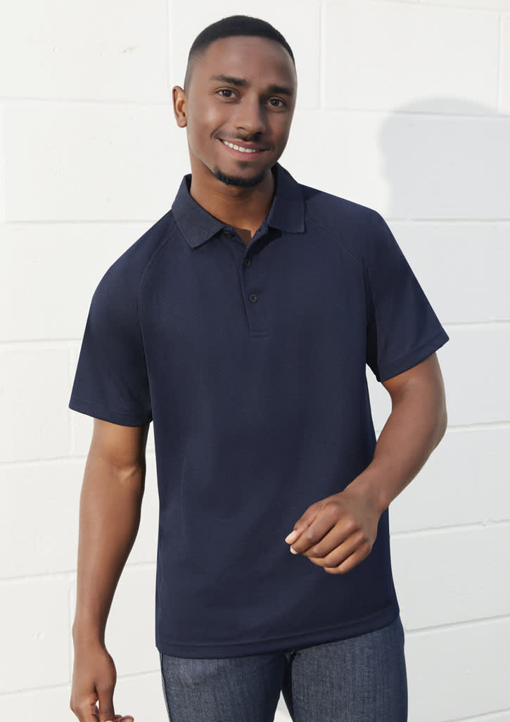 A navy short sleeve polo shirt with a collar and a logo, worn by a smiling man against a white wall.