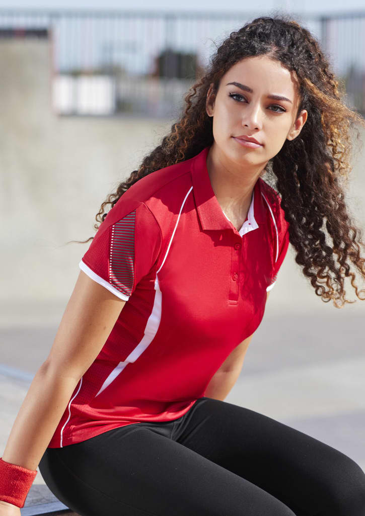 A red women's short sleeve polo shirt with white accents and a logo. The model is sitting outdoors.