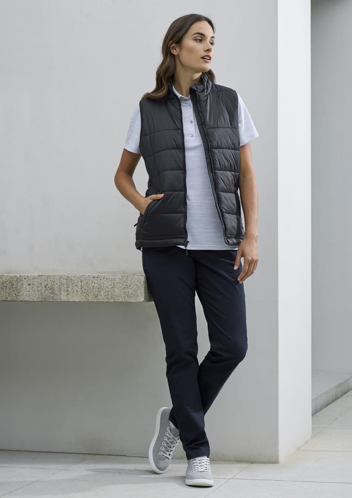 A woman wearing a grey short sleeve polo shirt, black padded vest, and navy pants, standing against a minimalist background.