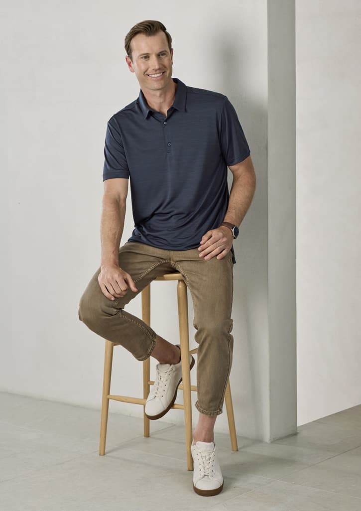 A man sits on a wooden stool, wearing a navy short sleeve polo and light brown pants with white shoes. The polo has a logo.