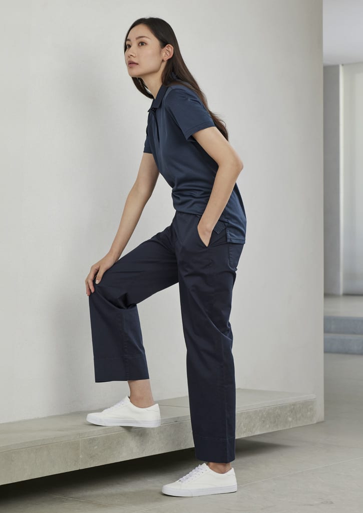 A woman is wearing a navy short sleeve polo shirt with matching dark trousers and white sneakers, standing against a wall.