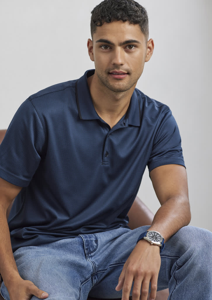A navy blue men's short sleeve polo shirt with a collar, featuring a logo, worn by a model sitting casually.