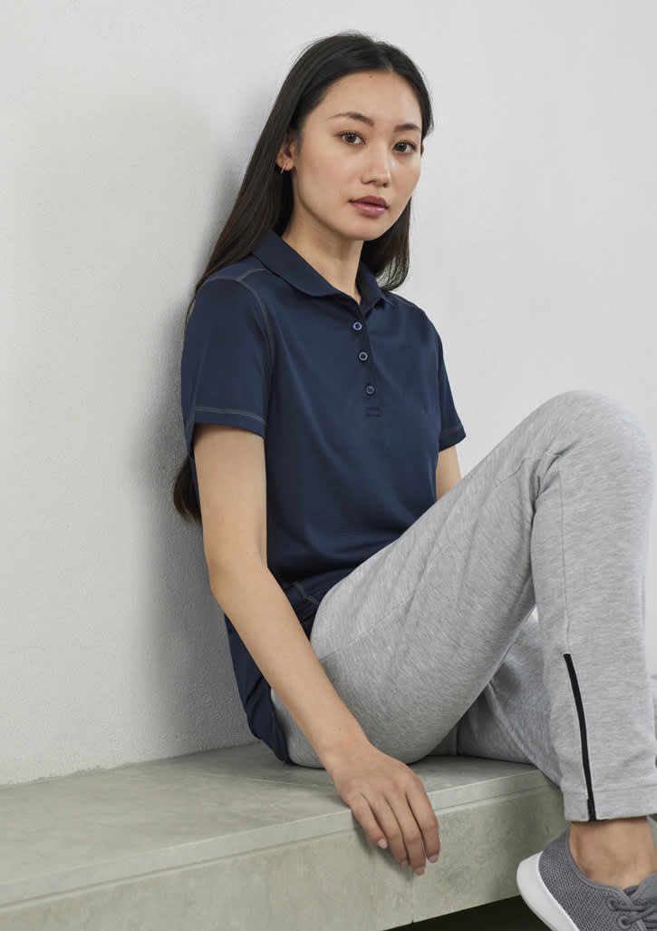 A woman wearing a navy short sleeve polo shirt sits on a bench, paired with grey athletic pants. The shirt features a logo.