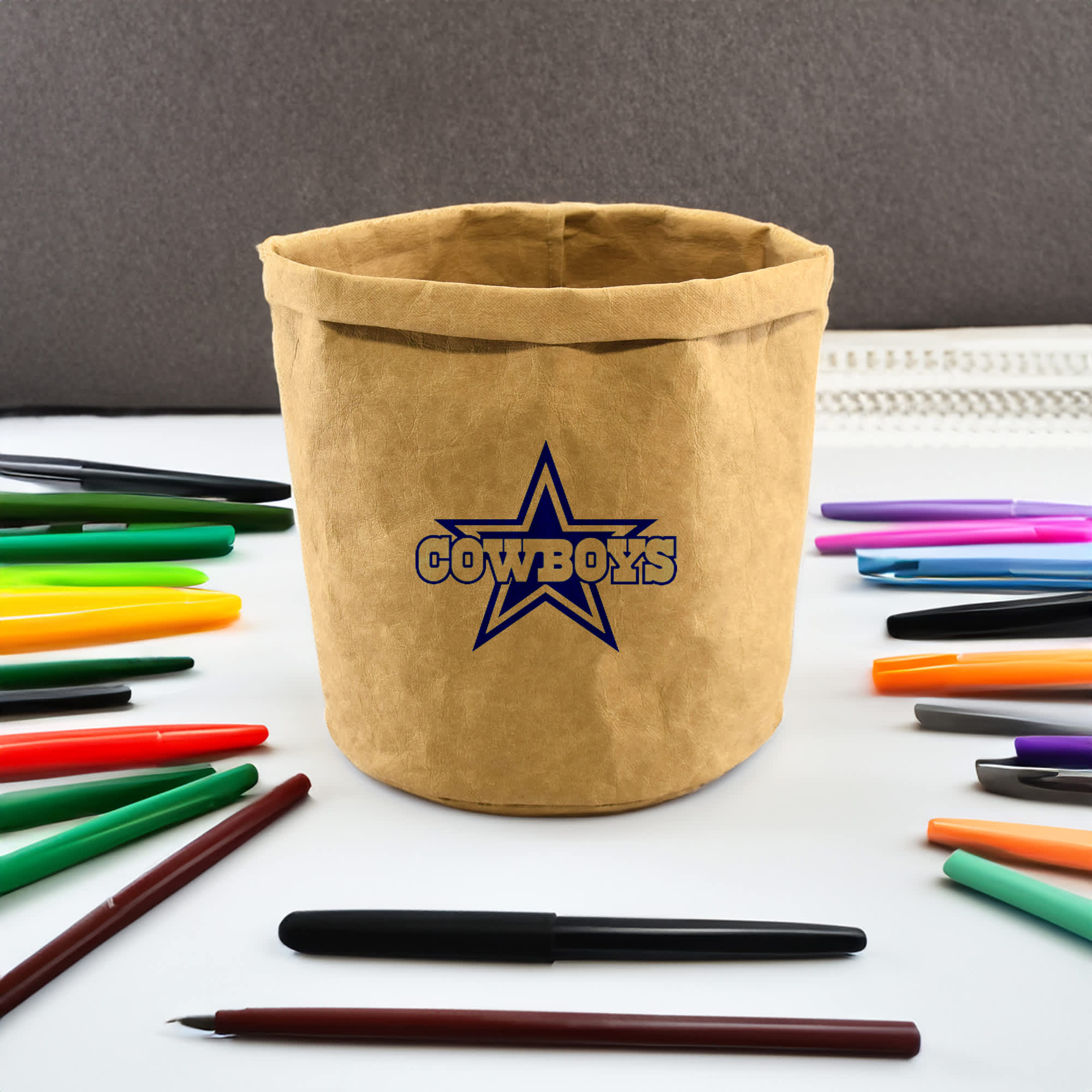A tan kraft paper storage bag with a logo, surrounded by various colourful pens.