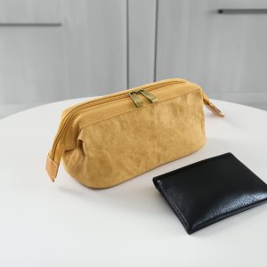 A yellow kraft paper cosmetic bag with a zip closure sits beside a black wallet on a white table. The bag has a logo.