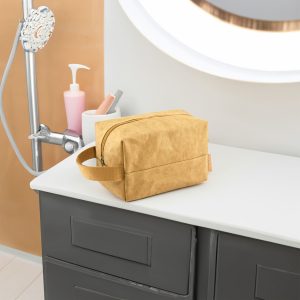 A kraft paper cosmetic bag in a warm yellow tone, placed on a bathroom countertop next to various toiletries.
