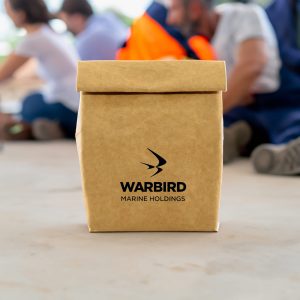 A kraft paper cooler bag in brown with a logo, placed on a surface in a casual setting.