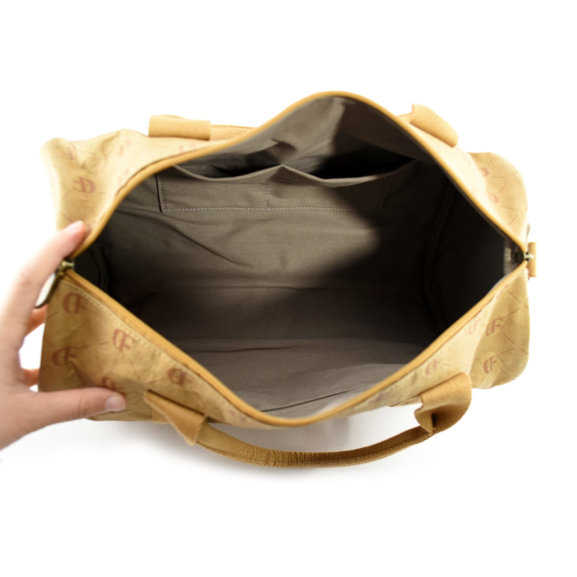 A hand holds a tan Manila Kraft paper duffle bag, showing its spacious, empty interior with a soft lining.