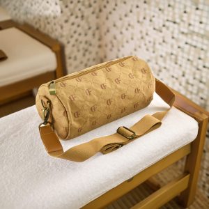 A cylindrical, ochre duffle bag with a logo, featuring a shoulder strap, placed on a white towel.