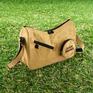 A tan Kraft paper sling bag with black accents and a round pocket featuring a logo. Set against green grass.