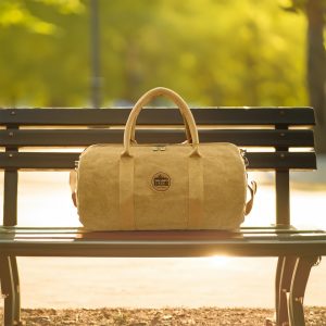 A kraft paper duffle bag in natural brown, featuring sturdy handles and a logo, resting on a park bench.
