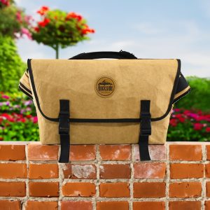 A tan kraft paper laptop messenger bag with black straps and a logo, set against a floral background.