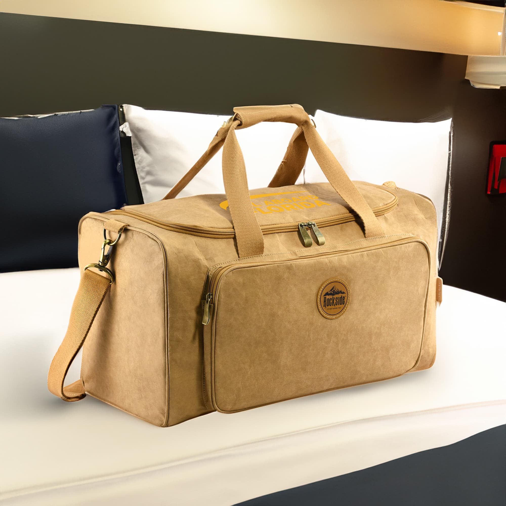 A kraft paper duffle bag in tan with handles and a detachable shoulder strap, placed on a bed.