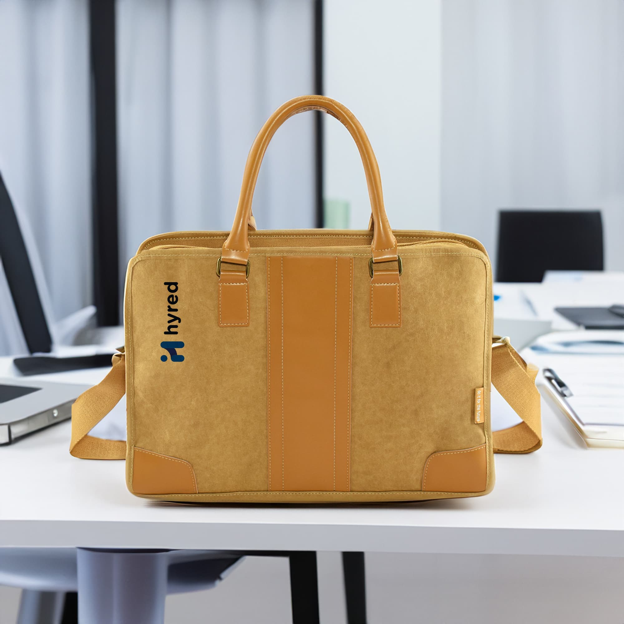 A yellow kraft paper satchel with tan accents and a logo, placed on a desk in an office setting.