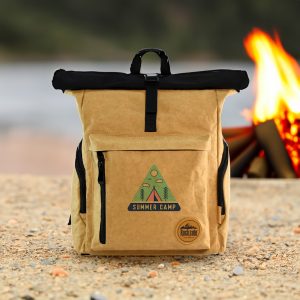 A tan kraft paper laptop backpack with a black top, featuring a logo and a zippered front pocket, set against a campfire backdrop.
