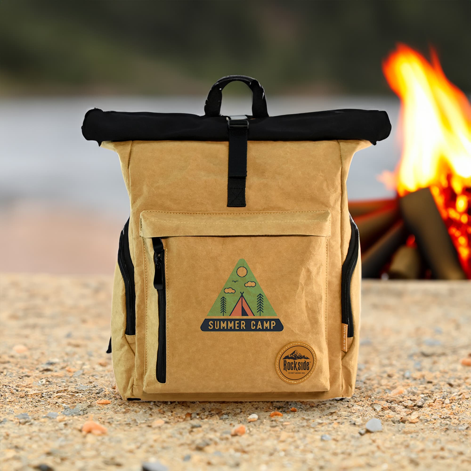 A tan kraft paper laptop backpack with a black top, featuring a logo and a zippered front pocket, set against a campfire backdrop.