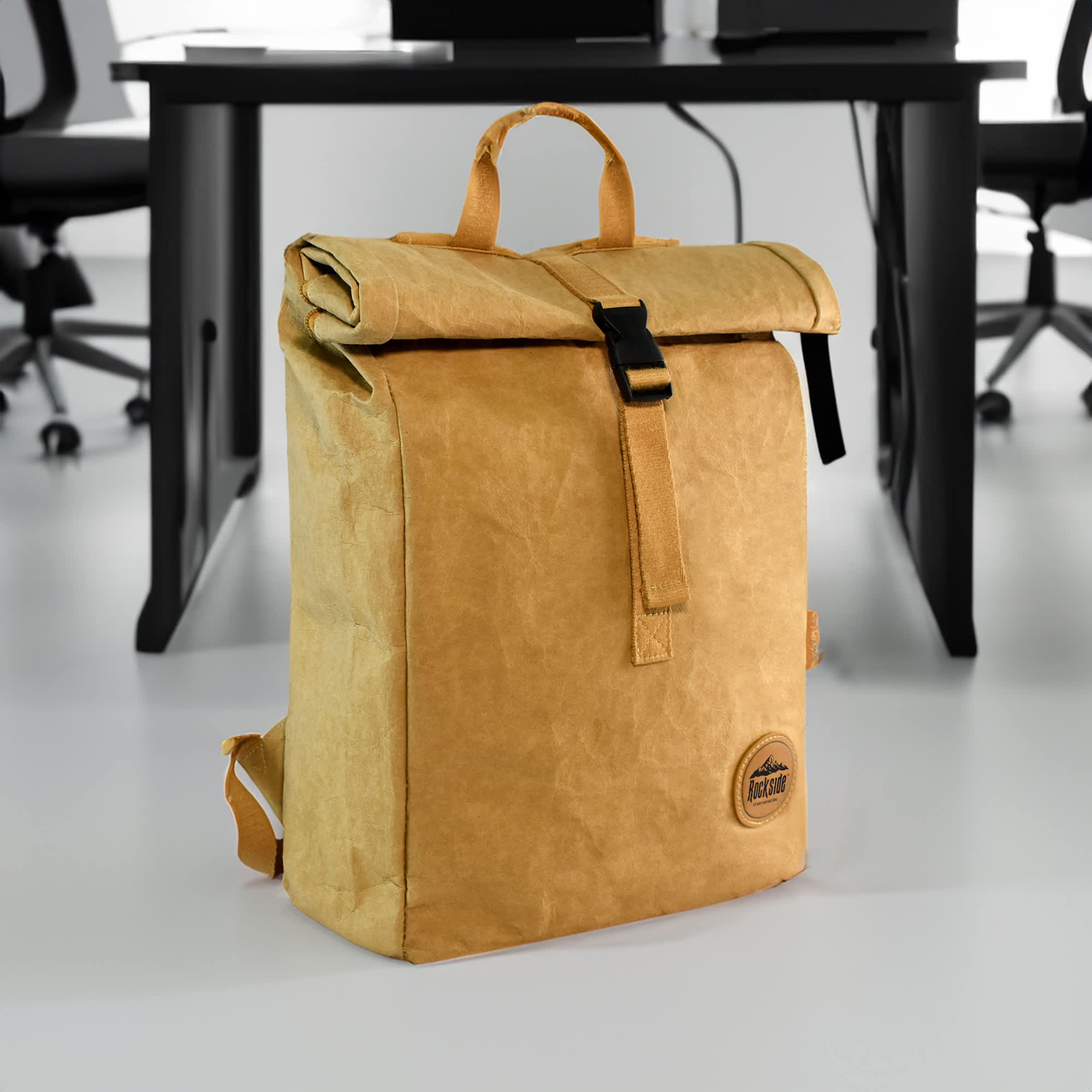 A tan kraft paper laptop backpack with a roll-top closure and a front buckle, featuring a logo.