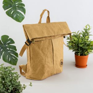 A yellow kraft paper laptop backpack with a roll-top closure, positioned beside potted plants.