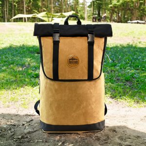 A tan and black backpack with a roll-top design and a logo, set against a natural outdoor background.